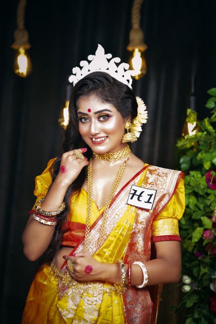 A young woman dressed in traditional Indian attire, wearing a yellow saree with red and gold accents, stands indoors with a dark backdrop illuminated by warm light bulbs. She has long, dark hair adorn