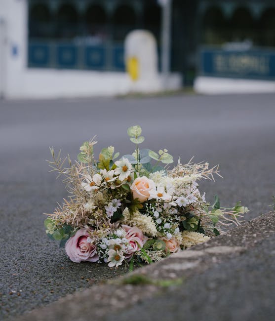 A bouquet of fresh flowers, including yellow sunflowers, white daisies, red gladiolus, and yellow lilies, wrapped in blue paper and arranged in a black plastic container, situated on the sidewalk next