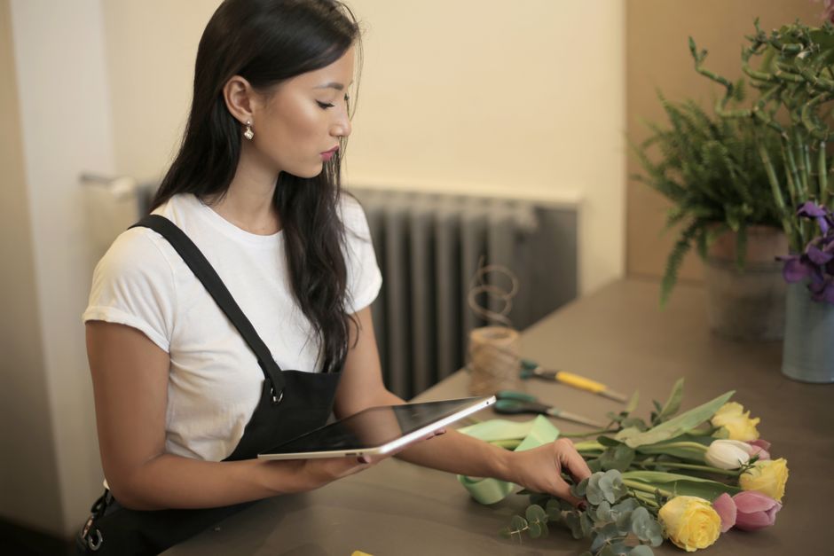 A flat-lay arrangement of fresh flower bouquets featuring pink tulips, yellow roses, and white blossoms with green foliage, placed around a tablet on a neutral background. The flowers are arranged wit