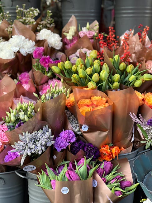 A person with short dark hair, wearing a white t-shirt, is carrying a large bouquet of fresh flowers wrapped in brown craft paper, positioned on their shoulder as they approach a florist shop with a b